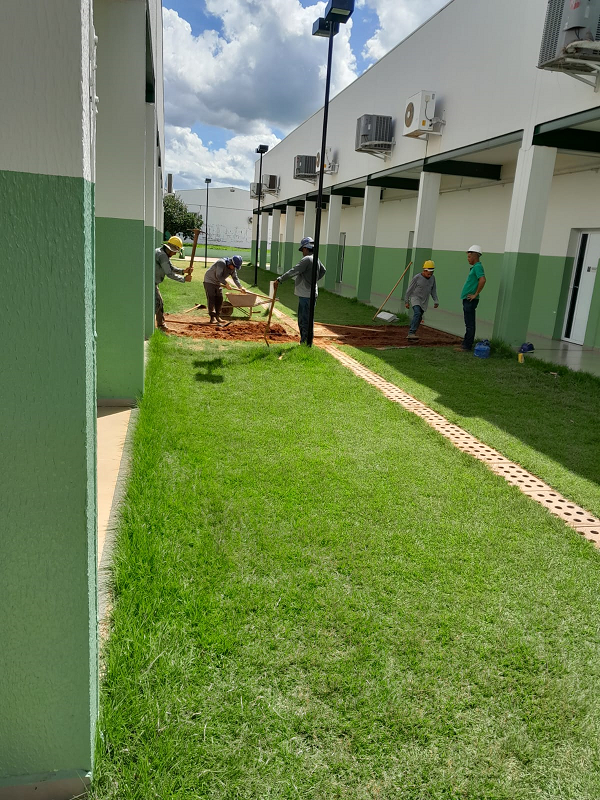 corredor de grama verde ao céu aberto, entre dois prédios, no segundo plano da foto, cinco homens estão trabalhando em uma passarela de terra que irá ligar os dois prédios