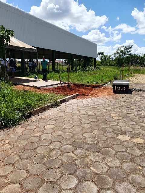 No primeiro plano aparece a rua de concreto, após alguns metros, do lado esquerdo, é possível ver a quadra coberta e entre a quadra e a rua, uma rampa de terra onde esta sendo construída a rampa de concreto.