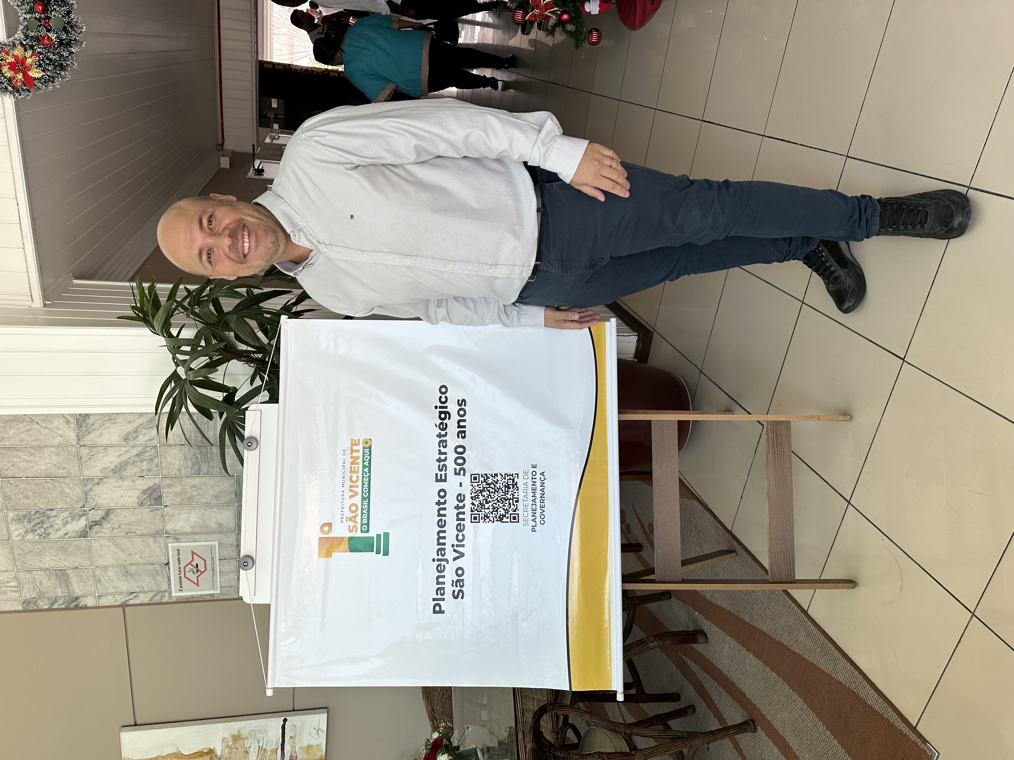 Foto. Professor Leandro Campelo posa para foto ao lado de um banner de fundo branco contendo a frase:  Planejamento estratégico. São Vicente 500 anos